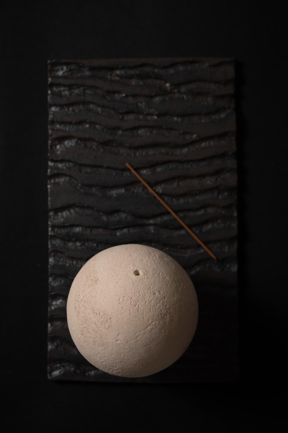 ceramic sphere on a ceramic black tray with wavy texture. incense stick lying on the tray.