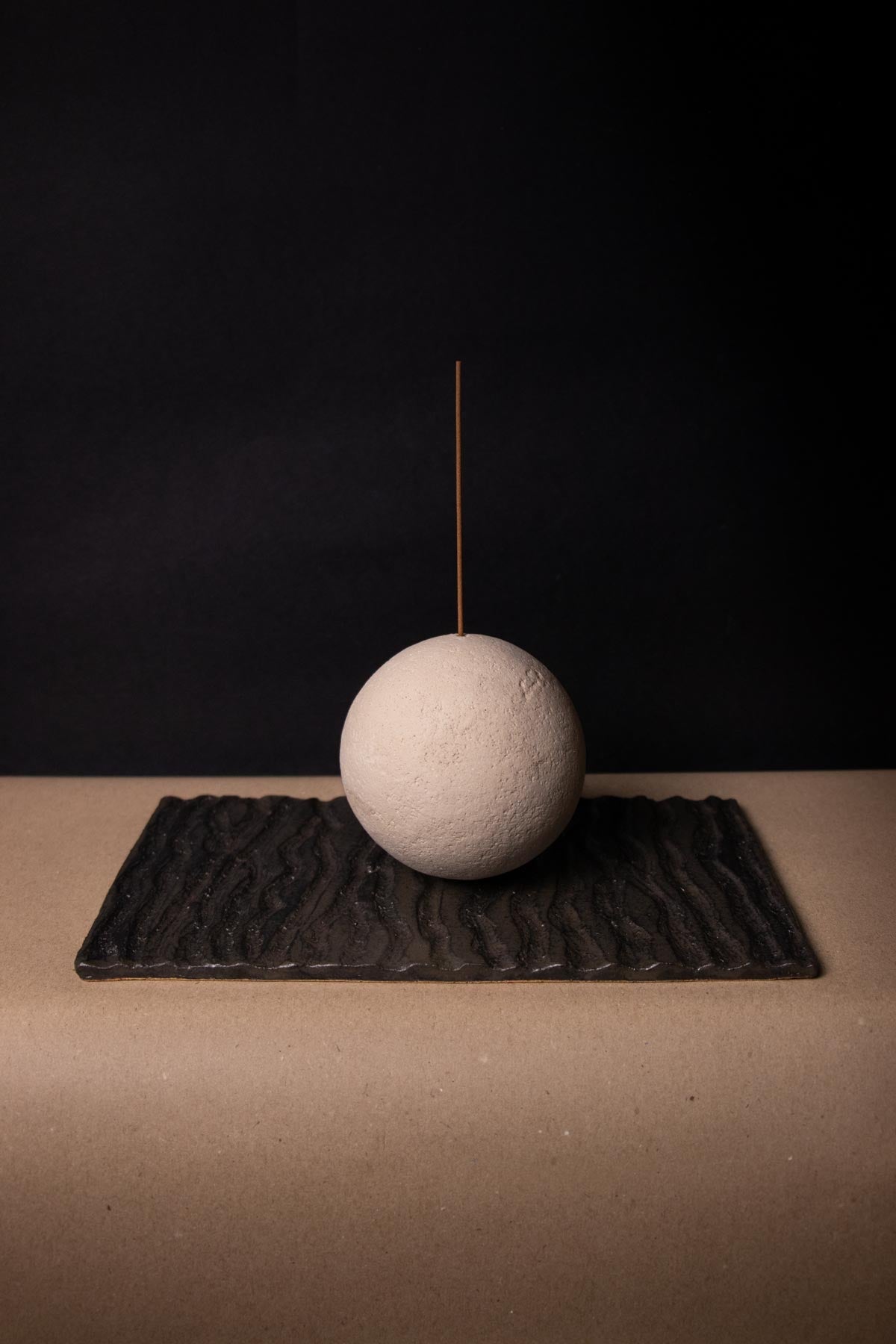 Incense burner inspired by the full moon. Ceramic sphere holding an incense stick on a wavy, black tray.