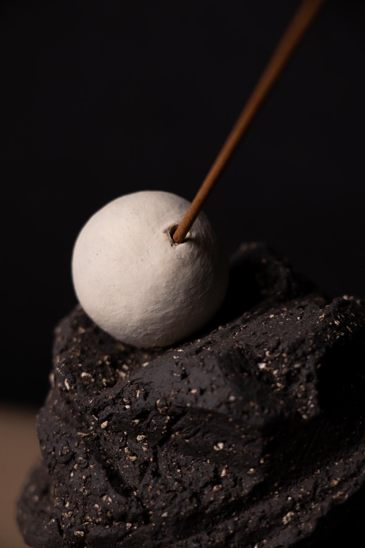 Close-up of incense stick in incense burner. White sphere. Black gritty stoneware. Ceramic incense holder.