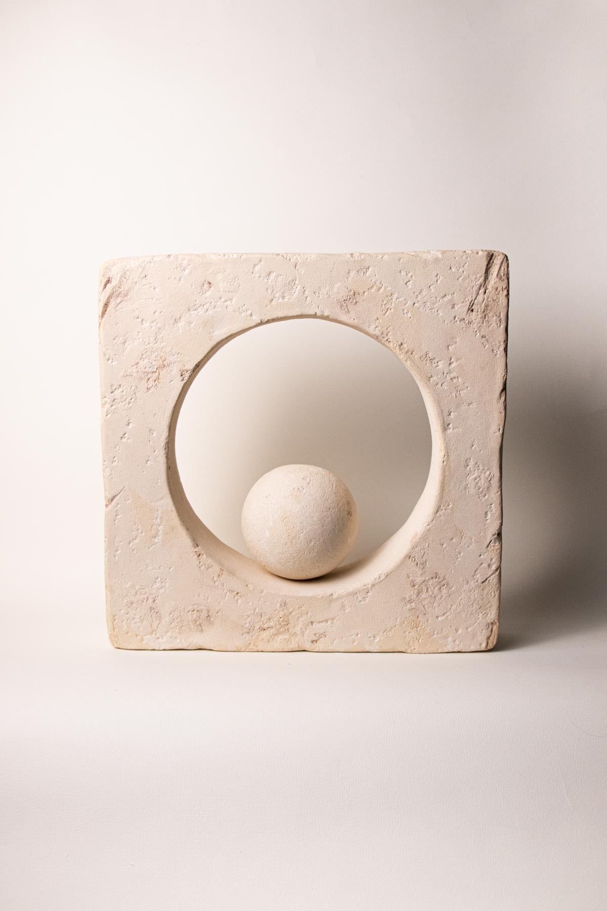 Ceramic square sculpture with a circular opening at the centre containing a sphere on an off-white background. Stoneware sculpture. Minimalist sculpture. Contemporary ceramics. Handmade ceramics. Organic texture. Cream and off-white colours. Neutral tones.