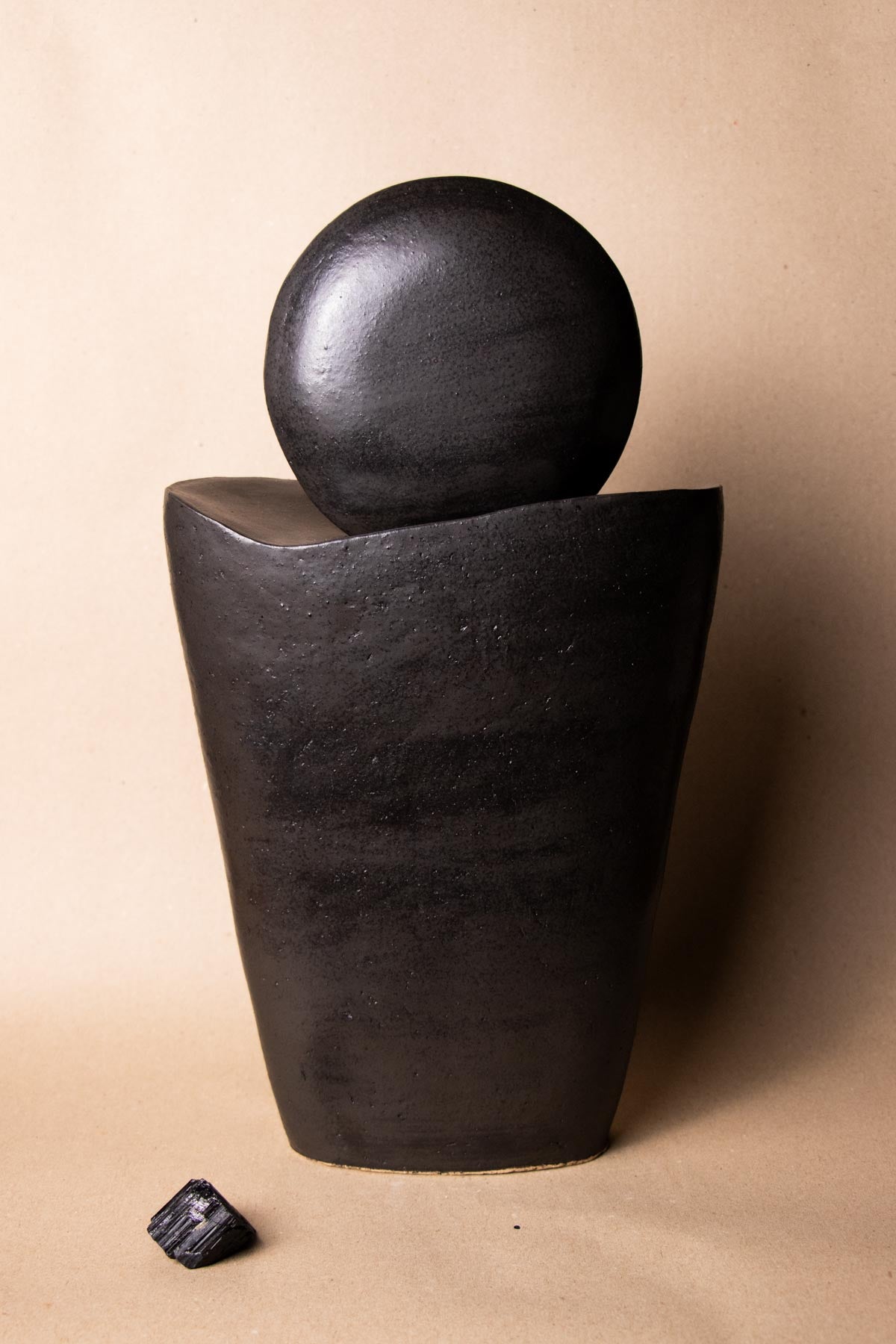 Black ceramic sculpture inspired by the moon. Minimalist stoneware sculpture. Minimalist decor. Decorative ceramics. In front of a plain white background and next to a piece of tourmaline crystal. New moon sculpture. Italian ceramics. Handmade in Italy.