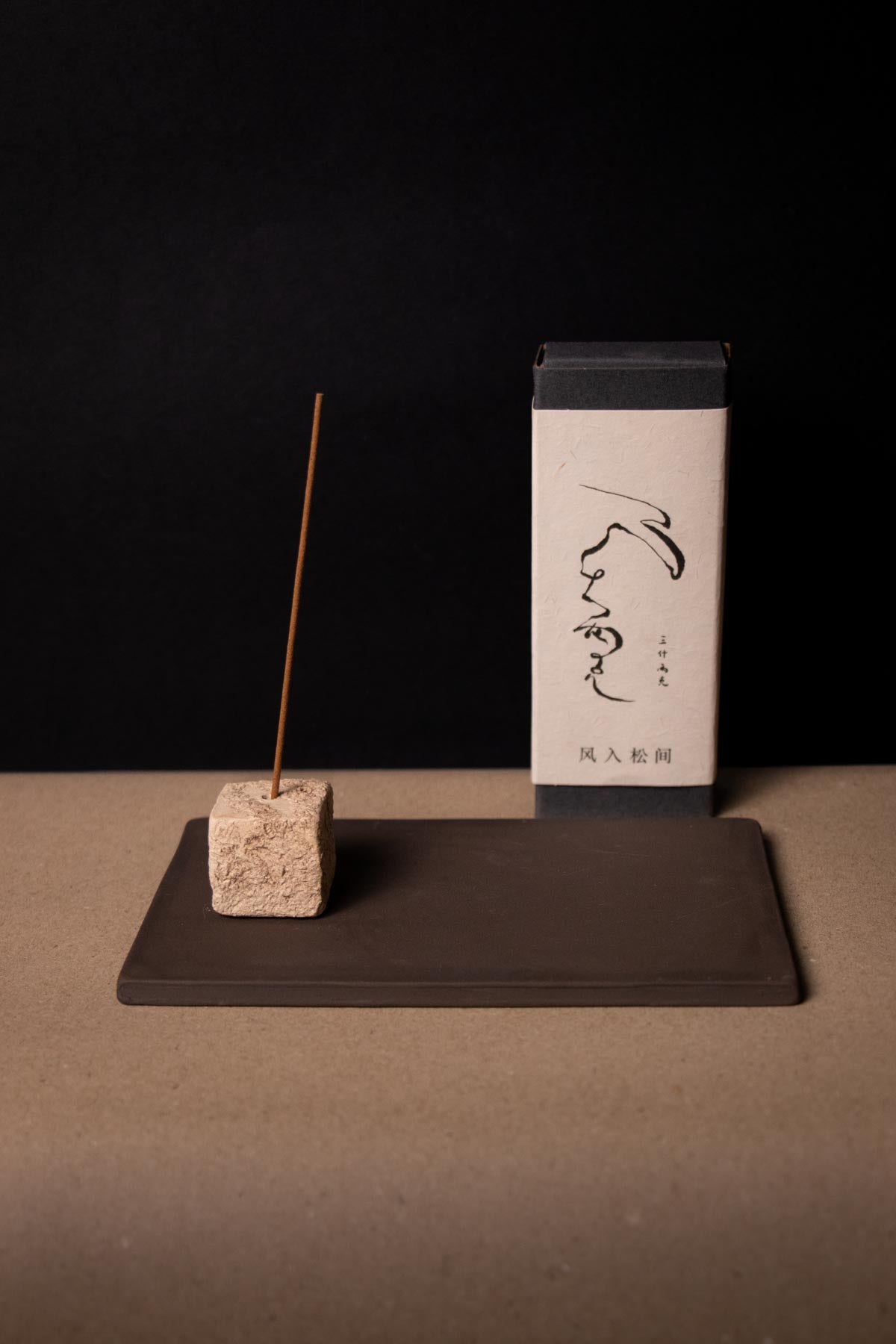 Incense stick and box on a dark surface with a black background. Artistic incense holder with brown base and a square, cream stick holder. Organic texture and wabisabi style