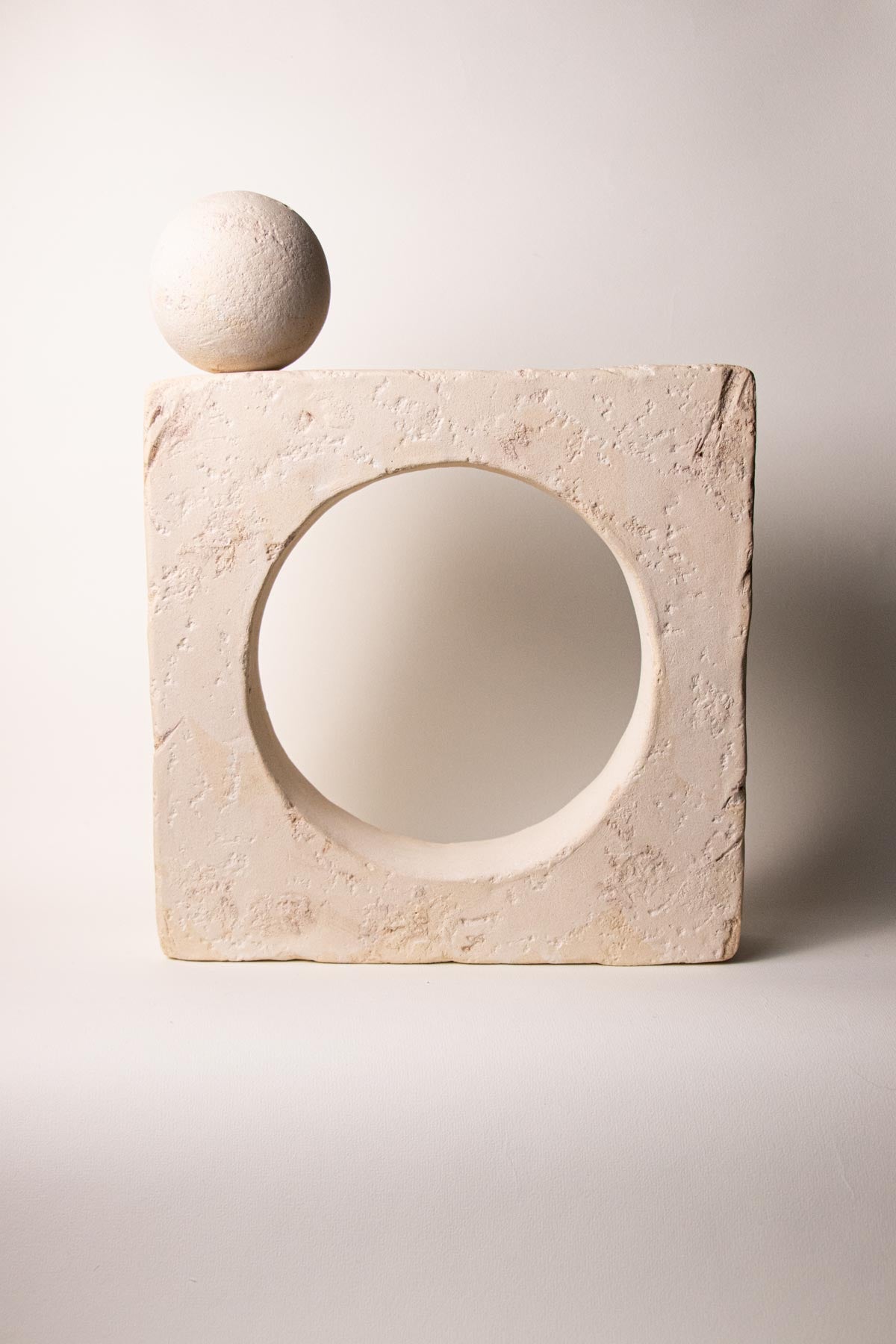 Ceramic square sculpture with a circular opening at the centre containing a sphere on an off-white background. Stoneware sculpture. Minimalist sculpture. Contemporary ceramics. Handmade ceramics. Organic texture. Cream and off-white colours. Neutral tones. 