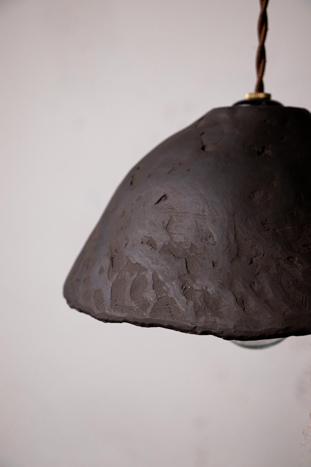 detail of Dark brown, ceramic pendant lamp with exposed bulb against a light gray wall. Minimalist design. Handmade ceramic lamp. Handmade ceramics. Stoneware lamp. 